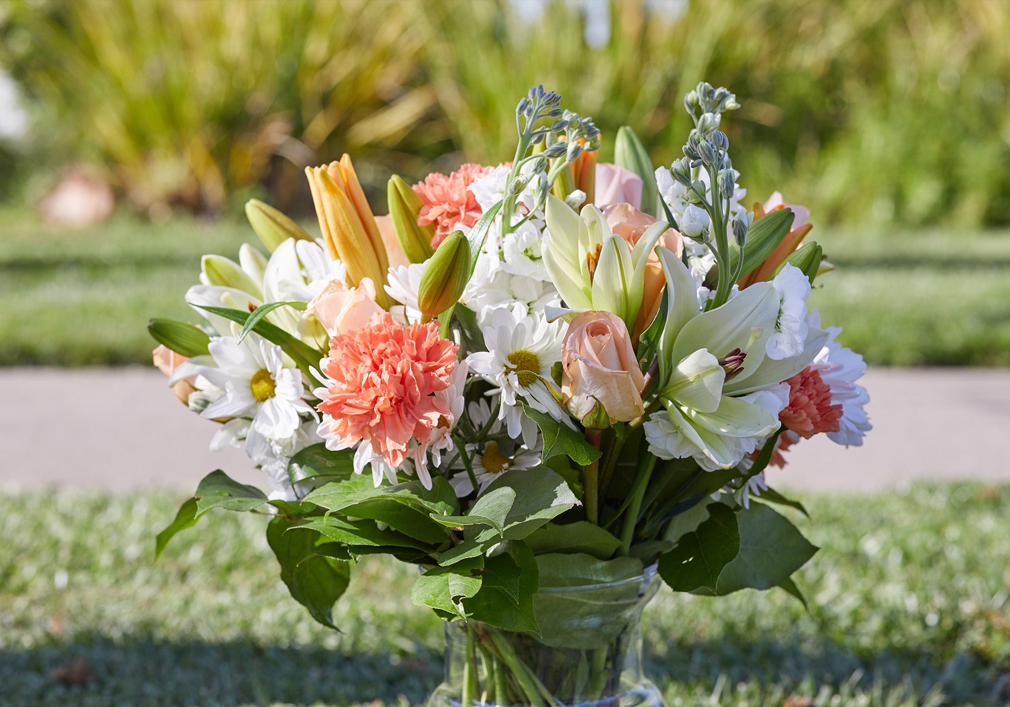 Bright floral arrangement featuring peach roses and carnations, white daisies, and white lilies, not quite in full bloom.